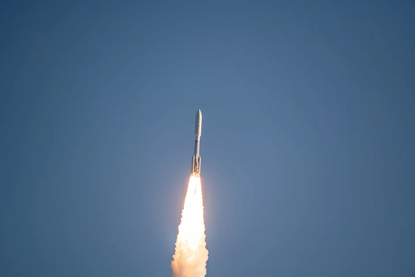 A white rocket climbs into a clear blue sky on a long column of flame and exhaust