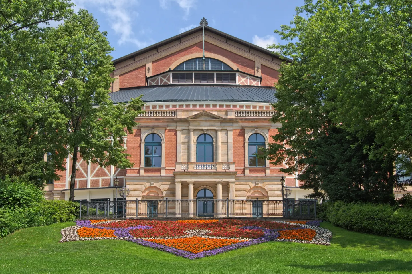 Red-brick front of the Bayreuth Festival Theatre with its columned balcony, seen across a lawn and flower beds