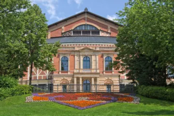 Red-brick front of the Bayreuth Festival Theatre with its columned balcony, seen across a lawn and flower beds