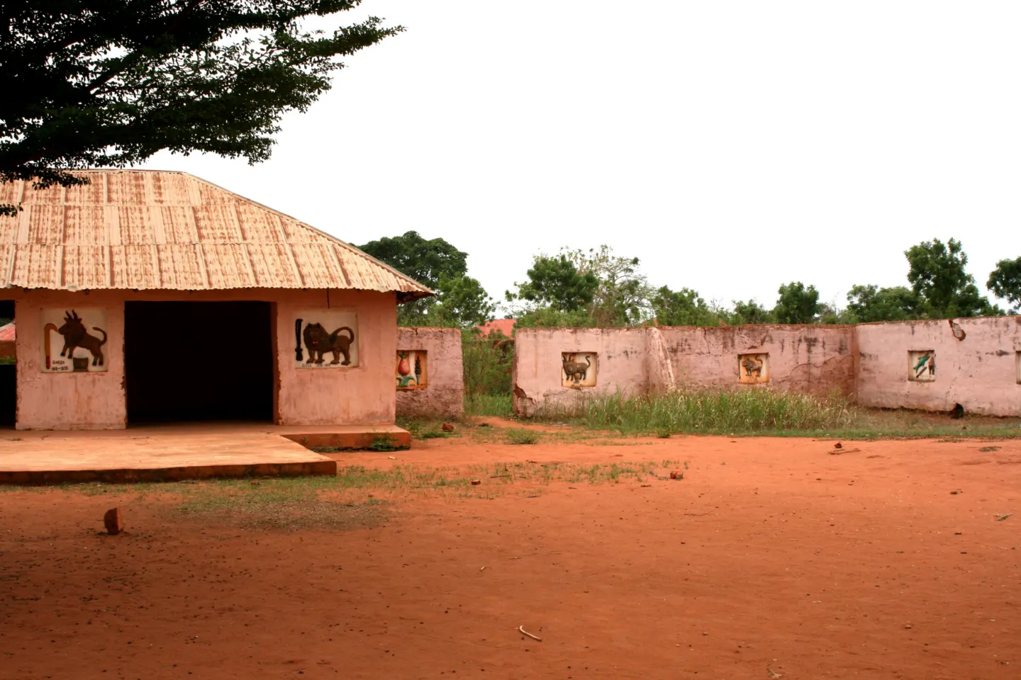 Low earthen palace building with a thatched roof and animal reliefs on its walls, beside a red-earth courtyard