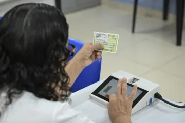 Seen from behind, a person holds a voter registration card while placing a finger on a biometric reader at a polling station