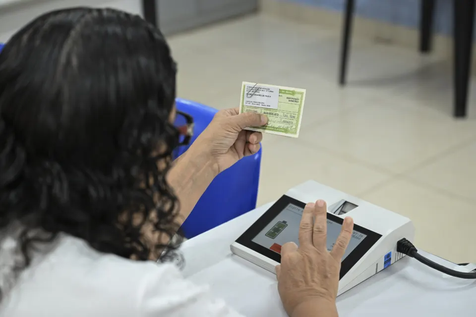 Seen from behind, a person holds a voter registration card while placing a finger on a biometric reader at a polling station