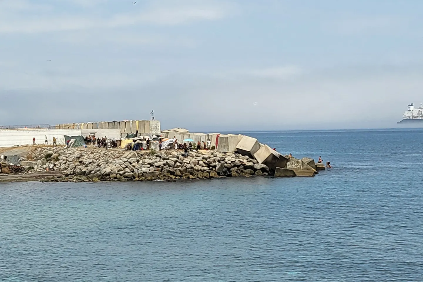 Rock breakwater in calm sea with makeshift shelters and small groups of people on it