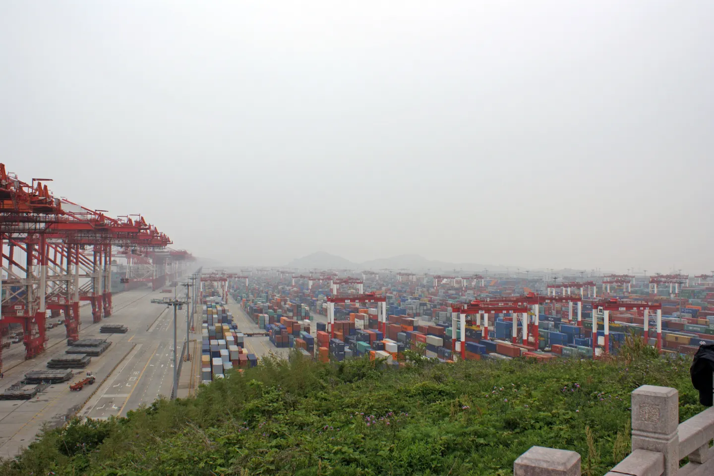 Rows of stacked shipping containers and red gantry cranes at a large seaport under a hazy sky