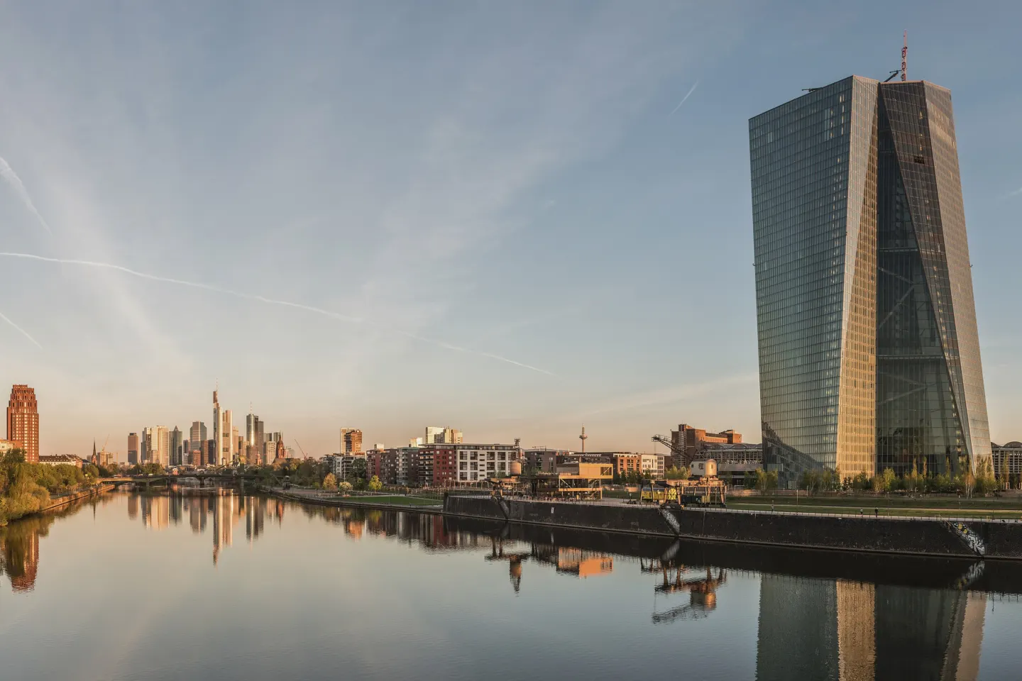 A twisting glass tower beside a calm river at dawn, with a cluster of skyscrapers in the distance