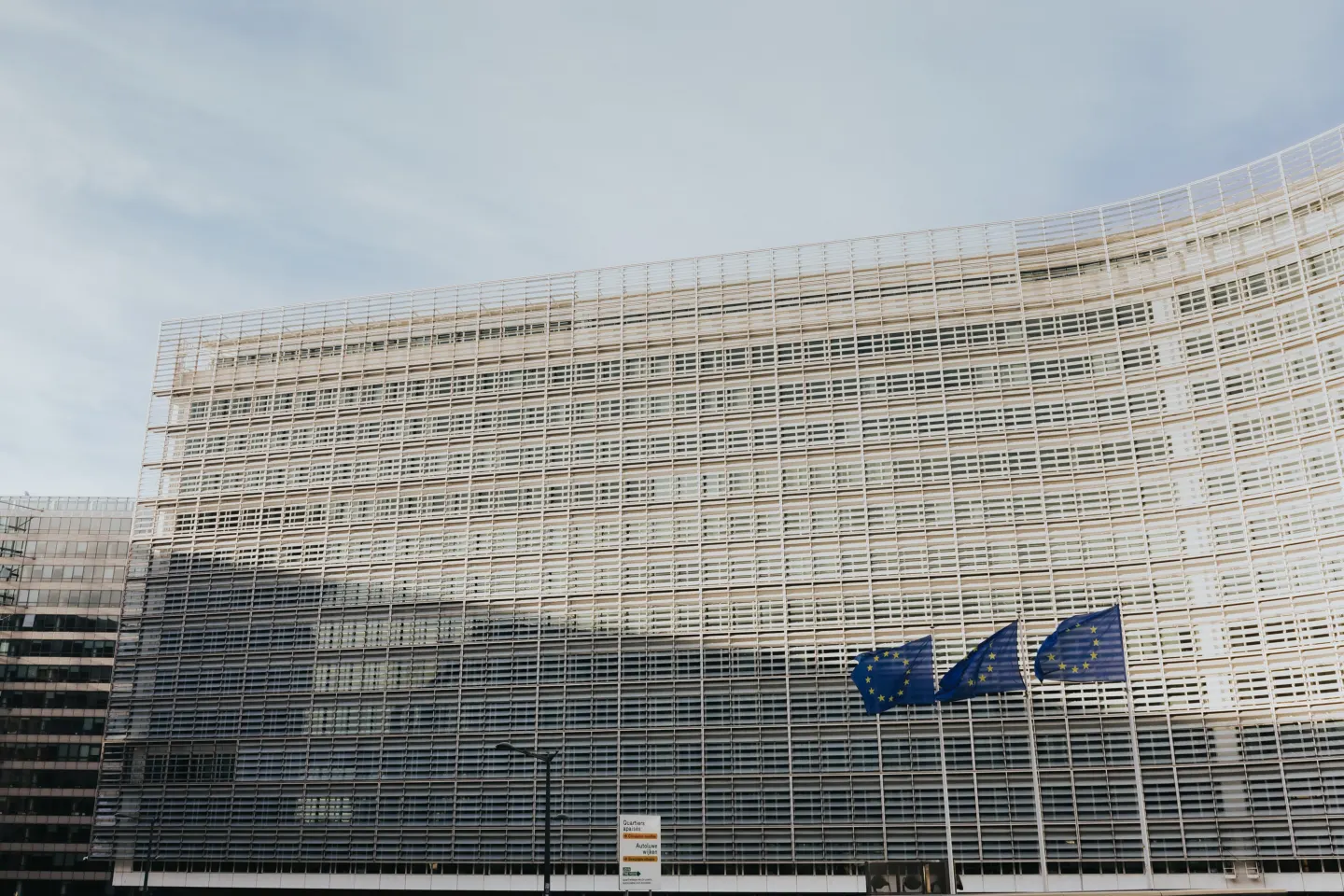 The curved, grid-patterned facade of a large office building with three European Union flags flying in front