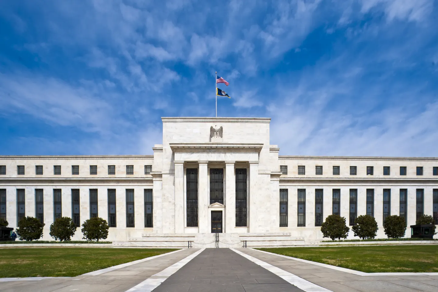 A wide white marble government building with tall columns at its entrance, a flag on the roof and a blue sky
