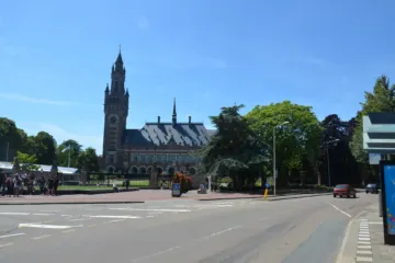 Brick palace with a tall clock tower behind trees and a road junction under a blue sky