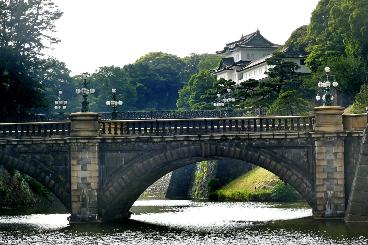 Stone double-arched bridge over a moat with a white palace keep among trees behind it