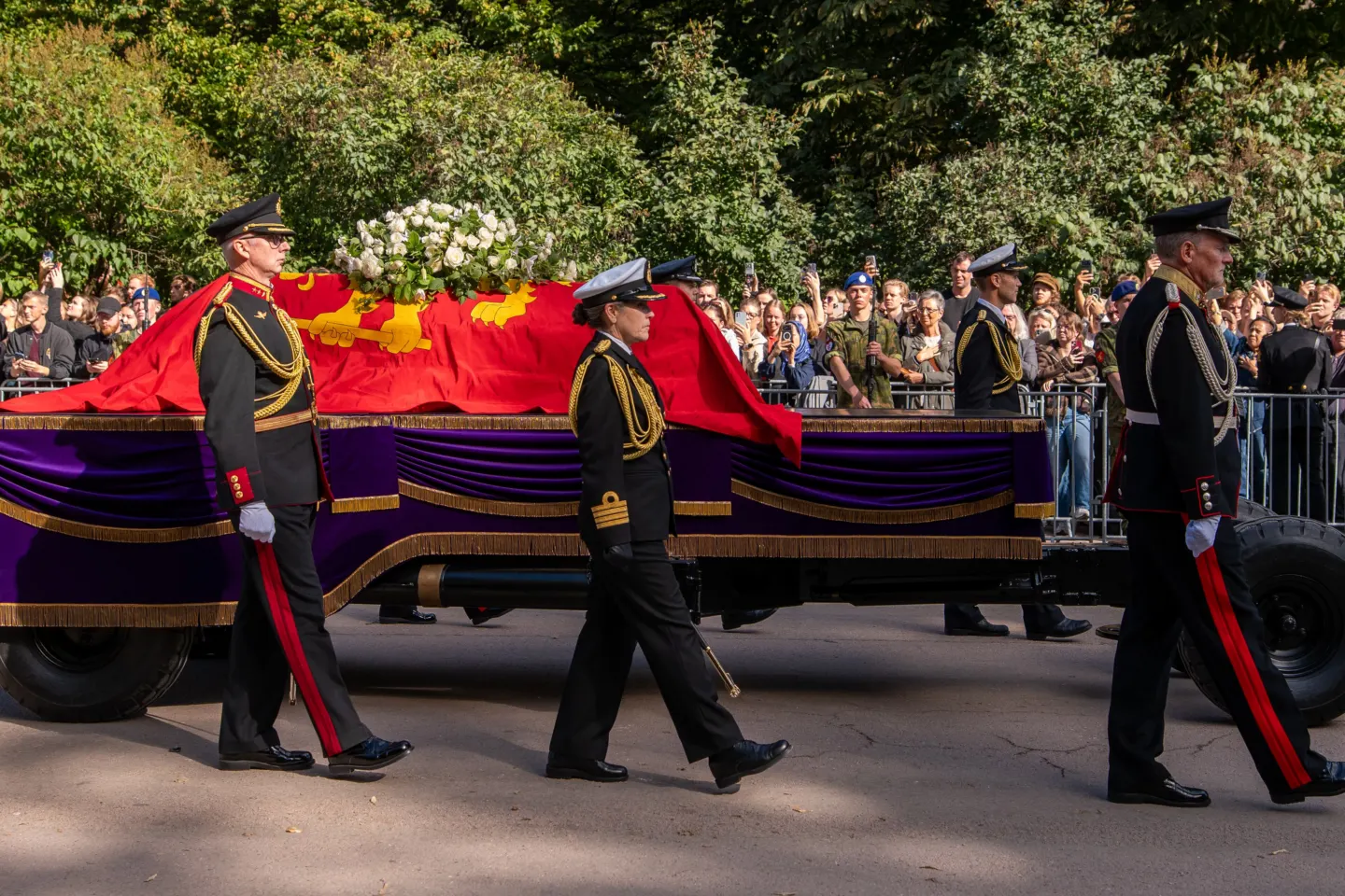 Uniformed officers walk beside a gun carriage bearing a coffin draped in a red royal standard, crowds behind barriers