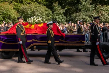 Uniformed officers walk beside a gun carriage bearing a coffin draped in a red royal standard, crowds behind barriers