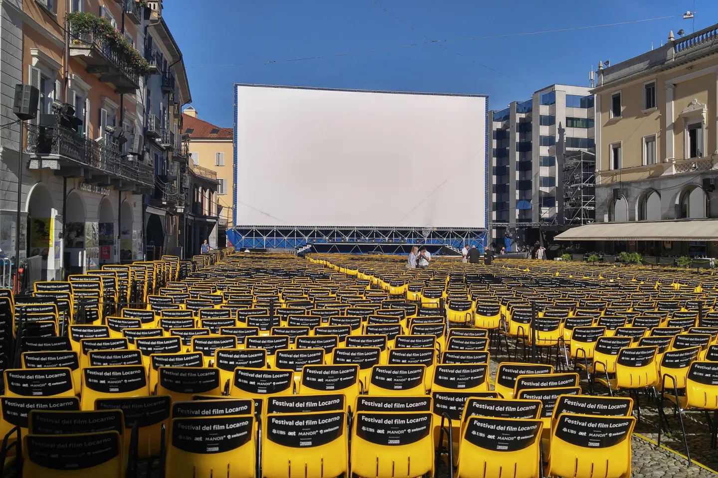 Rows of empty yellow and black chairs facing a very large outdoor cinema screen in a town square