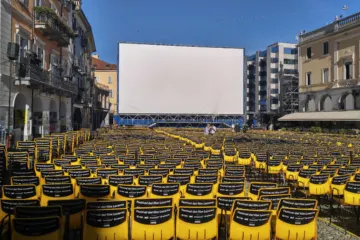 Rows of empty yellow and black chairs facing a very large outdoor cinema screen in a town square