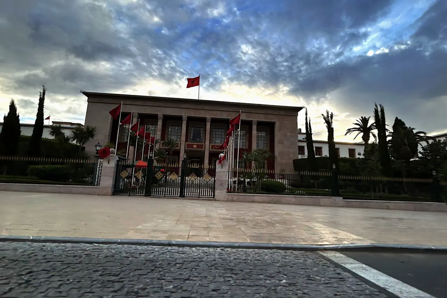 Colonnaded stone parliament building with a row of red Moroccan flags in front, under a cloudy evening sky