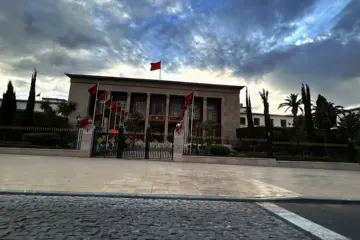 Colonnaded stone parliament building with a row of red Moroccan flags in front, under a cloudy evening sky