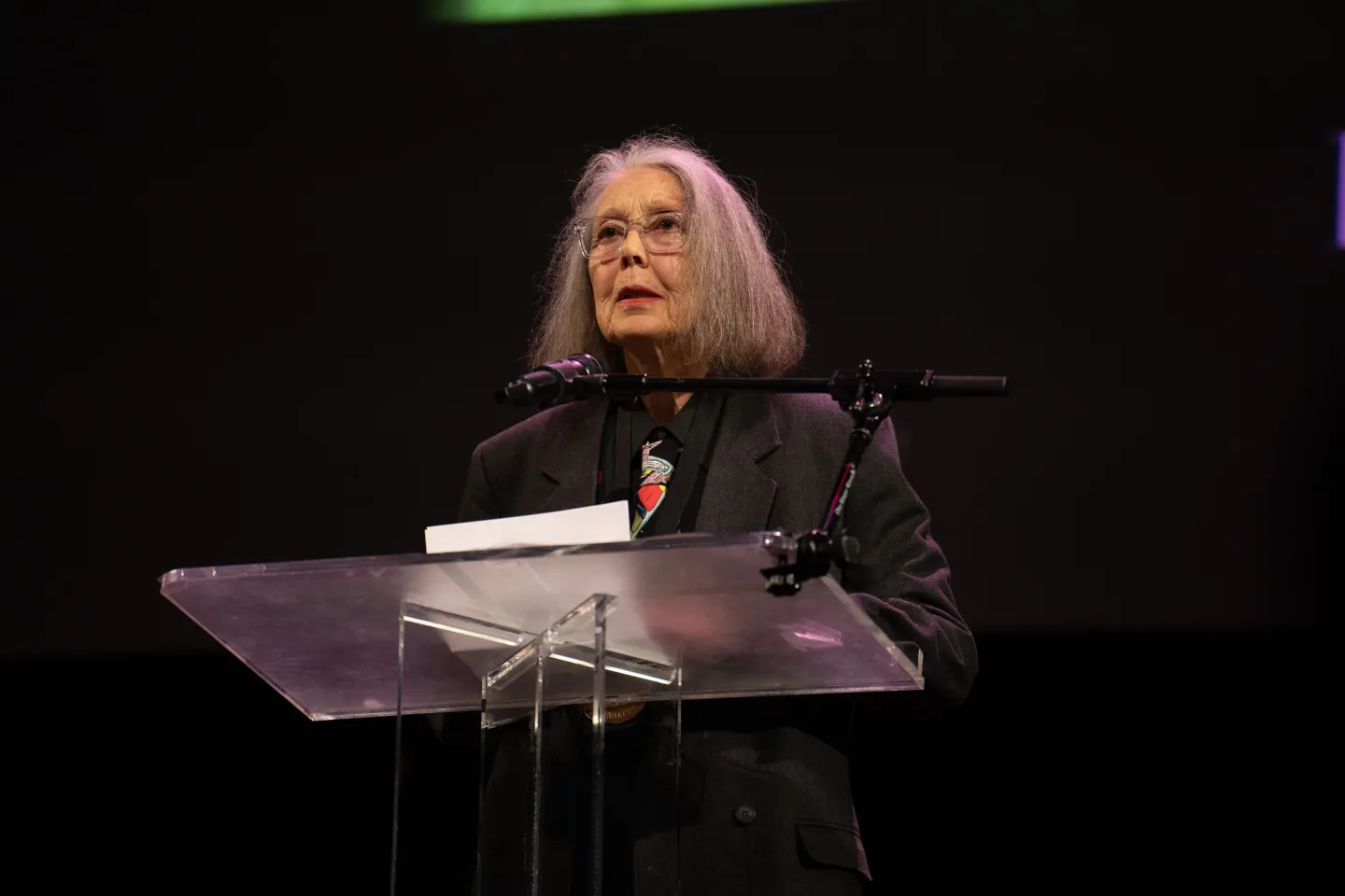 Anne Carson, a woman with long grey hair and glasses, speaking at a transparent lectern against a dark background