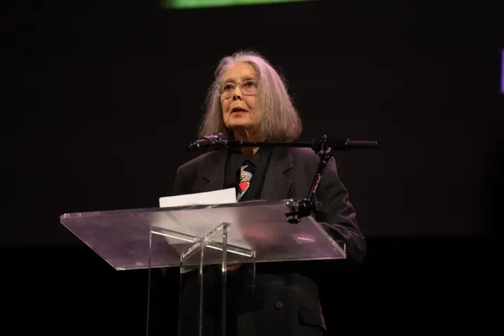 Anne Carson, a woman with long grey hair and glasses, speaking at a transparent lectern against a dark background