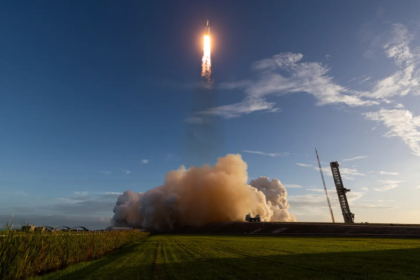 A rocket climbing on a bright flame above a large cloud of exhaust, with a launch tower and grass field in the foreground