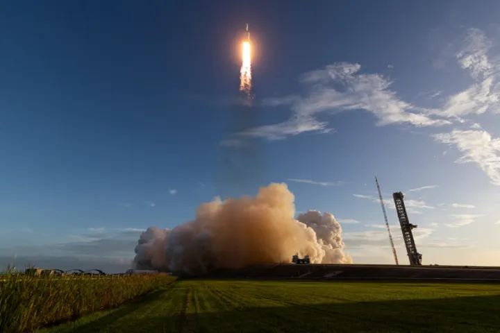 A rocket climbing on a bright flame above a large cloud of exhaust, with a launch tower and grass field in the foreground