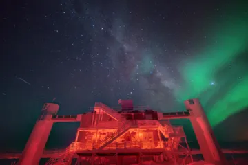 A red-lit building with two towers and outside stairs on the snow, under the Milky Way and a green aurora