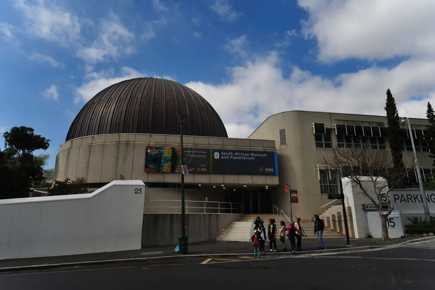 Concrete museum building with a large dark dome and a banner reading South African Museum and Planetarium; a few people on the steps