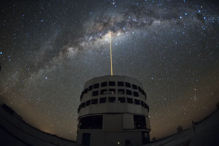 An orange laser beam rising from a telescope dome toward the bright band of the Milky Way at night