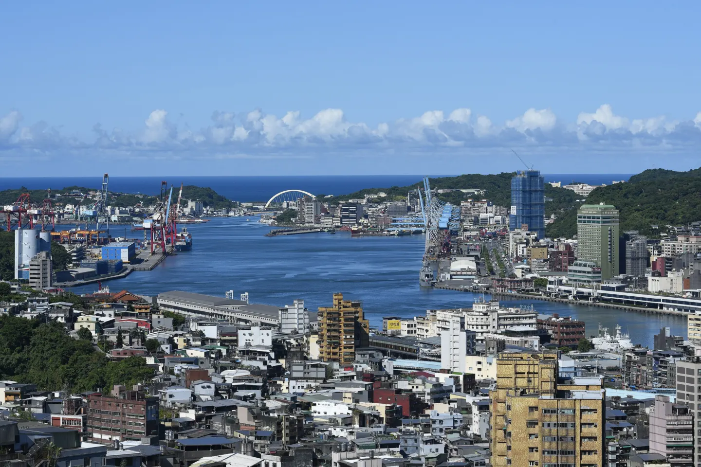 A harbour with container cranes and quays surrounded by dense city buildings and green hills under a blue sky