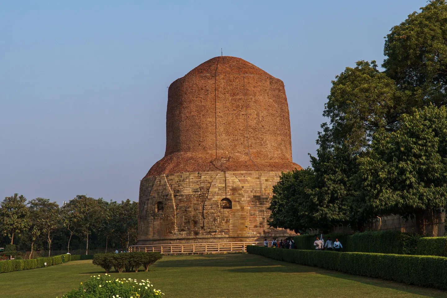 A tall cylindrical brick and stone stupa rising above lawns and low ruins under a clear sky