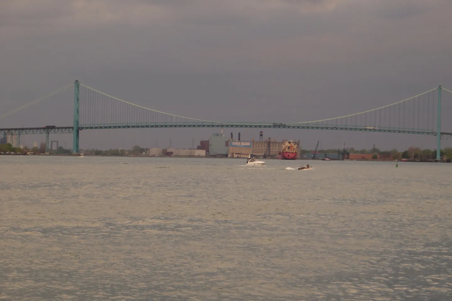 A long suspension bridge spanning a wide river under an overcast sky, seen from the water's edge