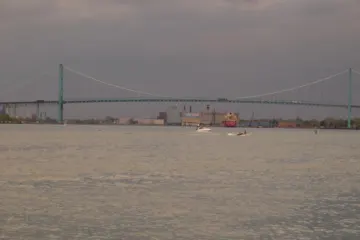 A long suspension bridge spanning a wide river under an overcast sky, seen from the water's edge