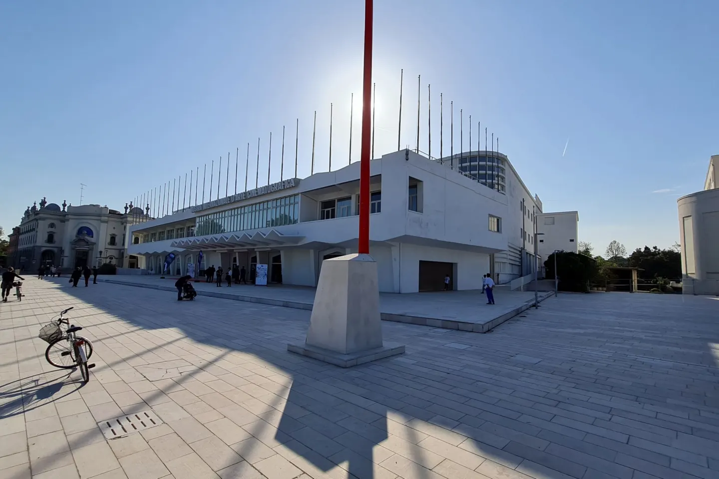Wide paved square in front of a low white modernist building topped with a row of flagpoles, in bright sunlight