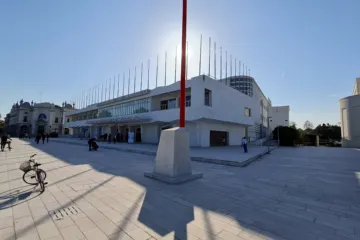 Wide paved square in front of a low white modernist building topped with a row of flagpoles, in bright sunlight