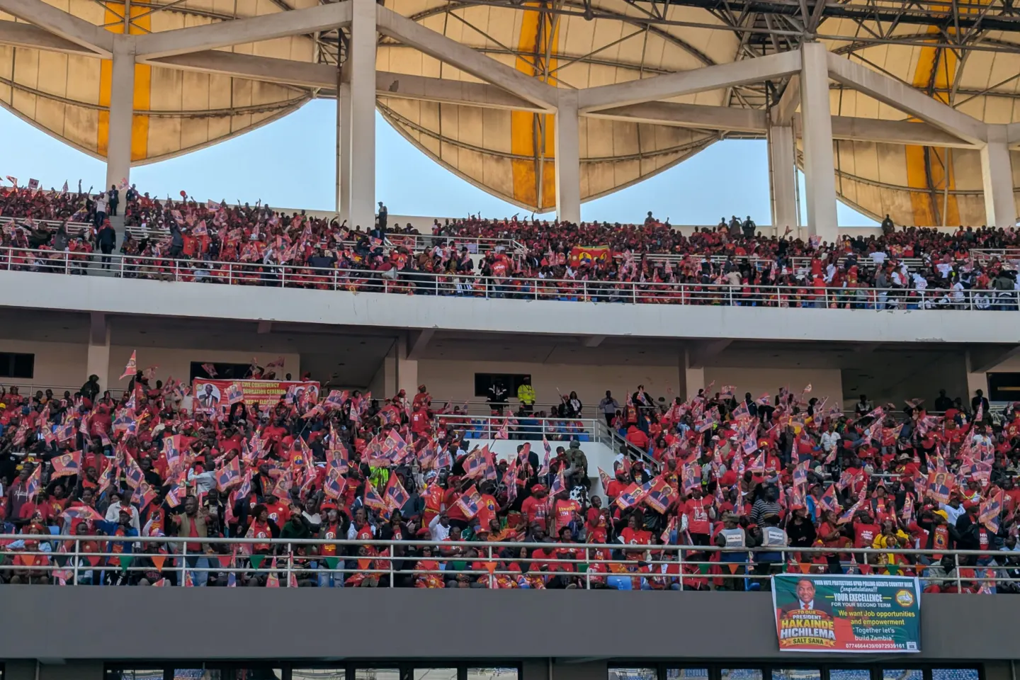 Packed stadium stands under a canopy roof, spectators in red waving small flags